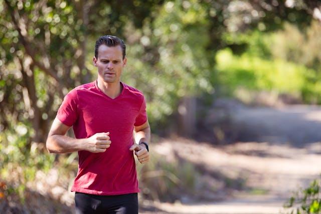 Homme brun avec tee-shirt rouge et short noir qui court sur un chemin bordé d'arbres