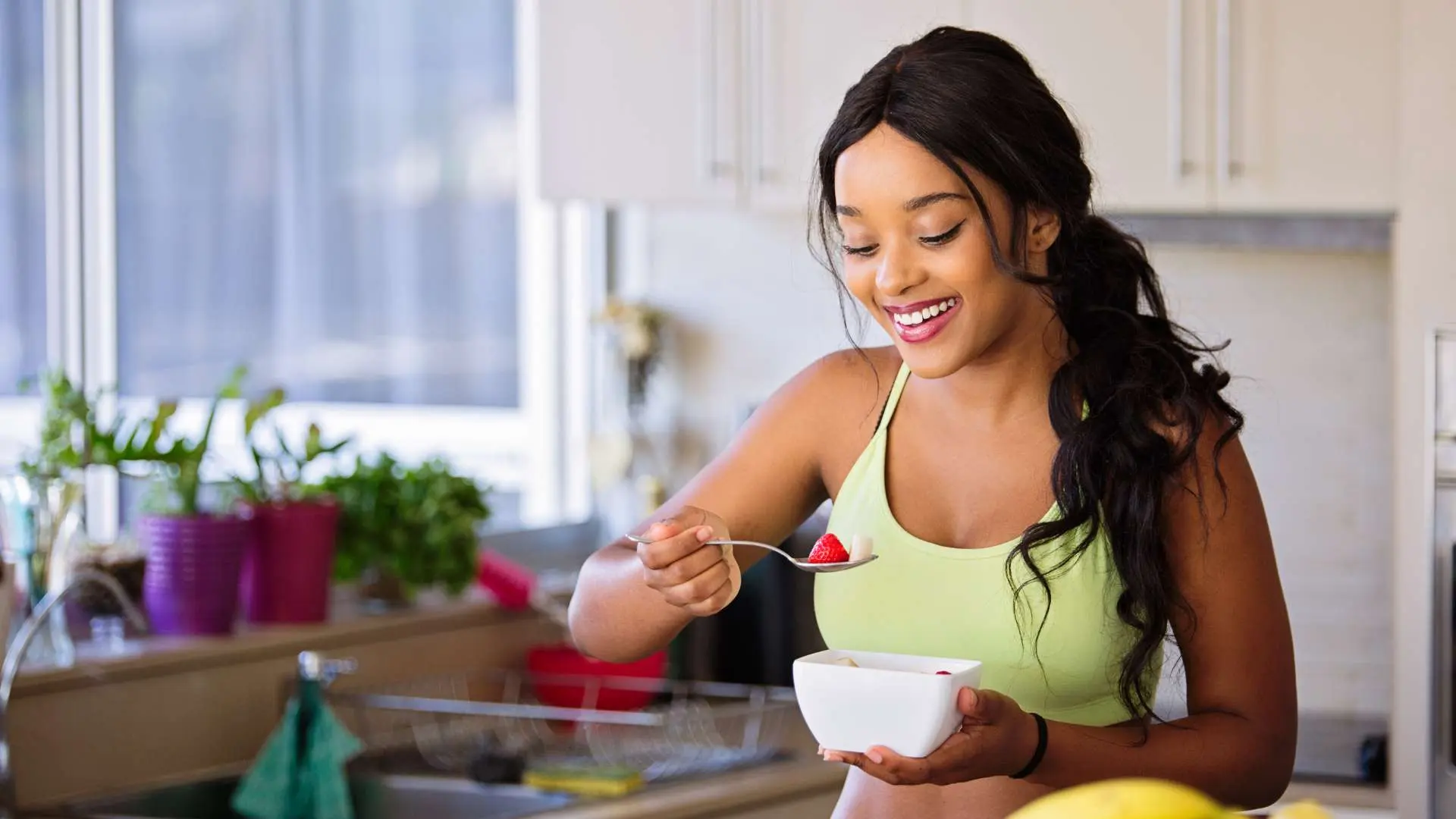 Jeune femme noire avec brassière verte dans une cuisine qui tient une cuillère avec fraise dedans et de l'autre main  un ramequin blanc
