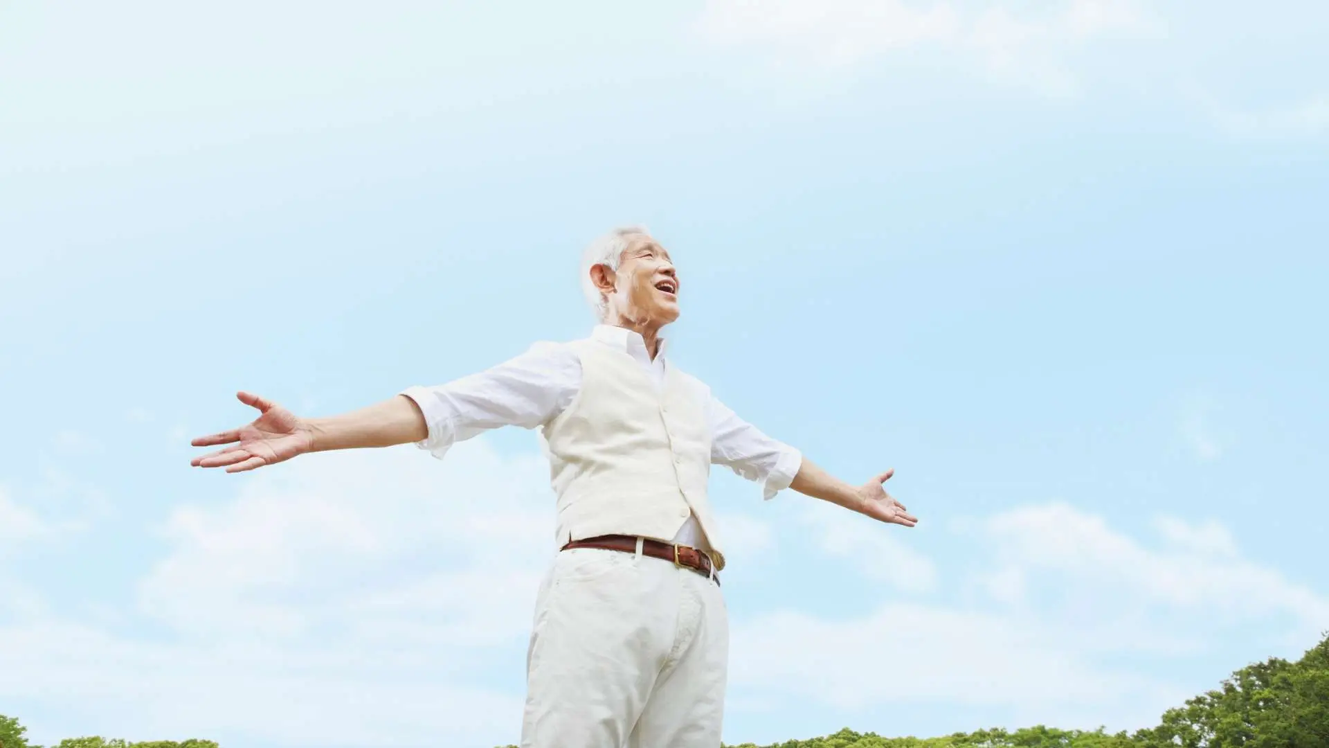 Homme senior habillé en blanc souriant bras étendus avec ciel bleu en arrière-plan et cimes d'arbres vertes
