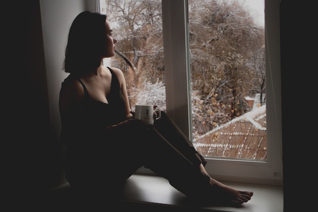 fille triste assise sur un rebord de fenêtre avec une tasse de thé qui regarde dehors