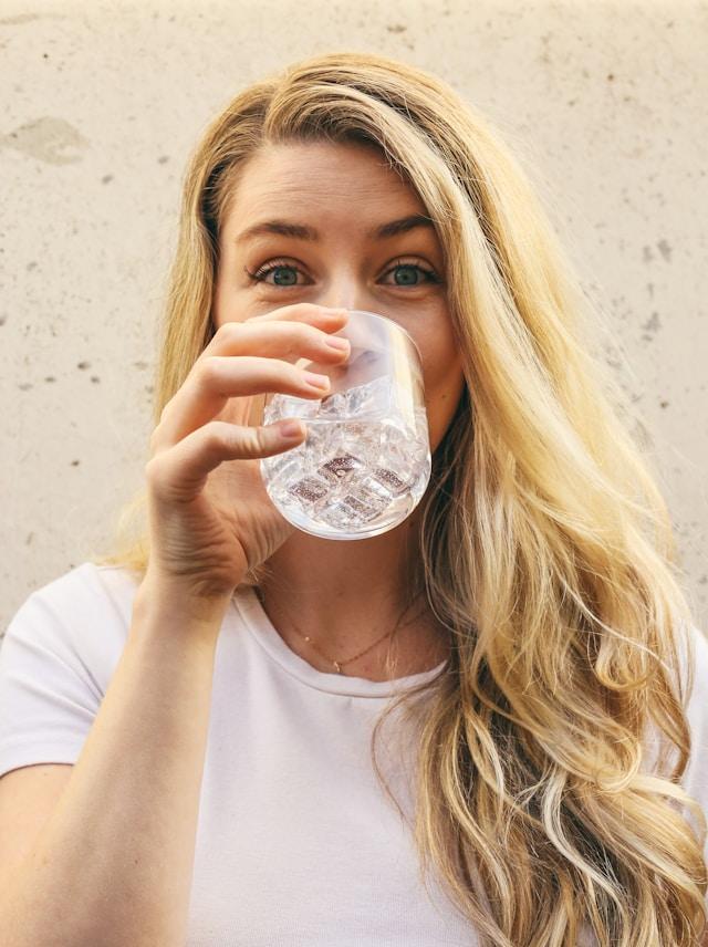 femme blonde cheveux longs avec tee-shirt blanc manches courtes qui sourit et boir un verre d'eau