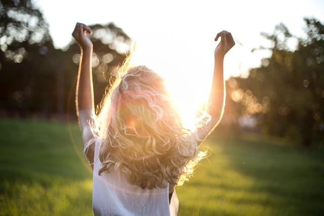 Femme blonde cheveux longs et ondulés de dos qui lève les bras face au soleil