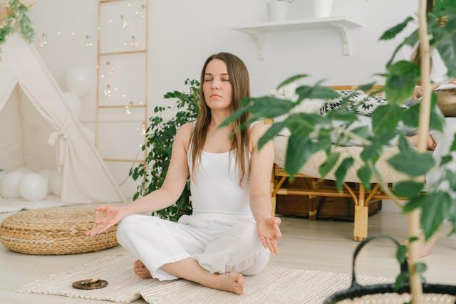 Femme brune cheveux longs qui médite en tailleur dans un salon avec des plantes vertes et décoration blanche et en bois