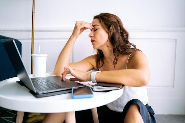 Femme cheveux longs châtains avec caraco blanc assise devant table blanche basse avec un ordinateur, un gobelet blanc avec paille, un téléphone portable à coque bleue qui se tient le front avec une main les yeux fermés