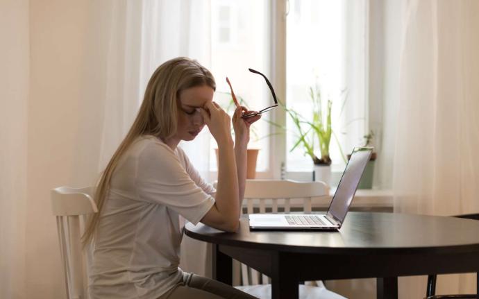 Jeune femme cheveux longs blonds et tee-shirt blanc devant ordinateur qui tient ses lunettes dans une main et de l'autre main se tient le front les yeux fermés