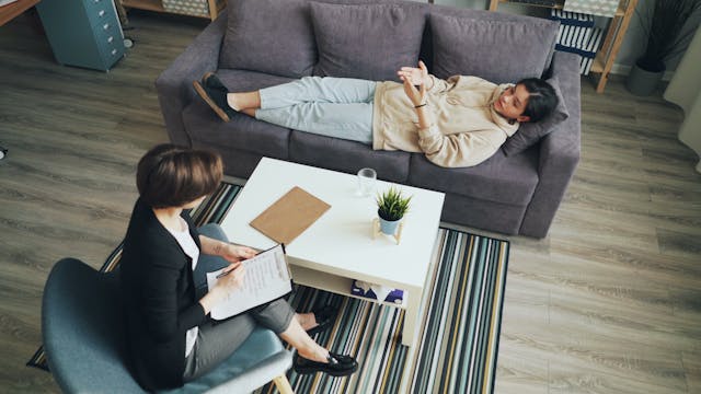 Jeune femme allongée sur un divan avec une psychologue en face assise dans un fauteuil bleu et une table blanches les séparant