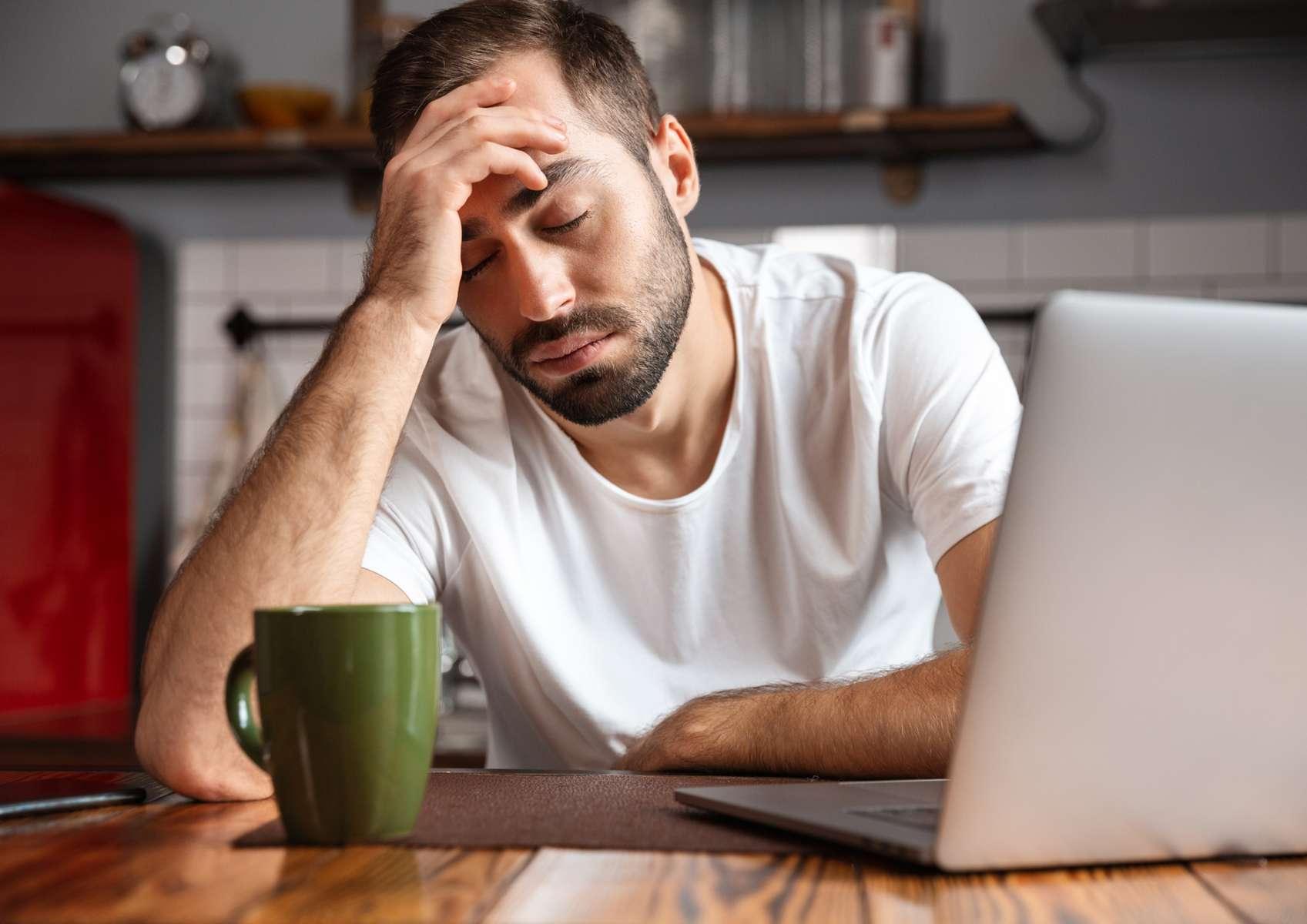 Homme brun avec tee-shirt blanc fatiguée les yeux fermés assis sur une chaise dans une cuisine avec à côté de lui une tasse verte et devant lui un ordinateur ouvert