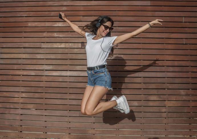 Jeune femme brune lunettes de soleil, portable à la main, baskets et tee-shirt bma,cs qui saute devant un mur en bois
