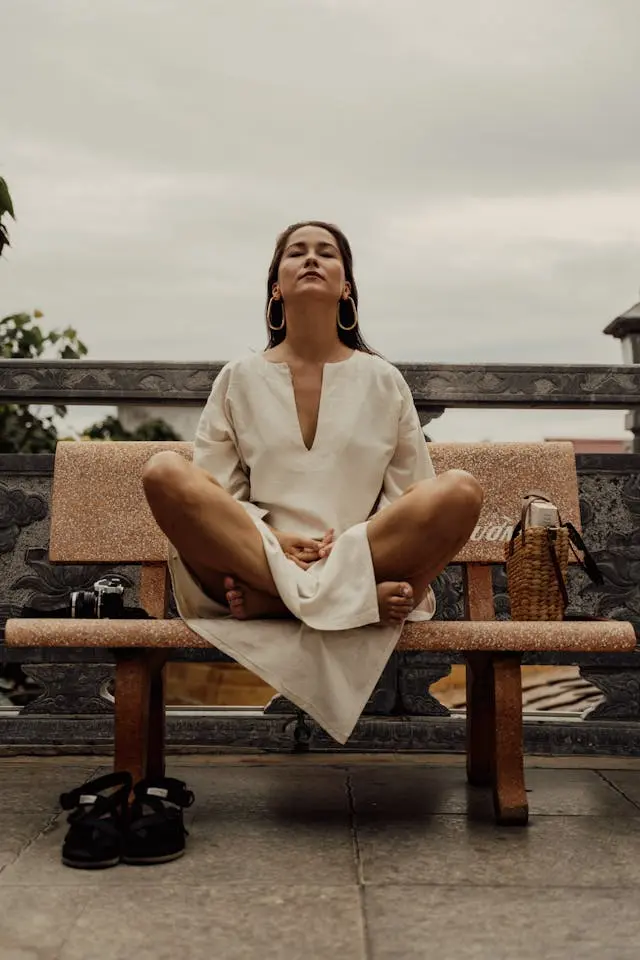 Femme  en tenue blanche qui médite sur un banc avec un sac à côté d'elle et ses chaussures à terre