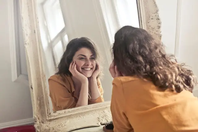Jeune femme bruine avec chemise orange qui sourit devant un miroir les mains en coupe sur ses joues