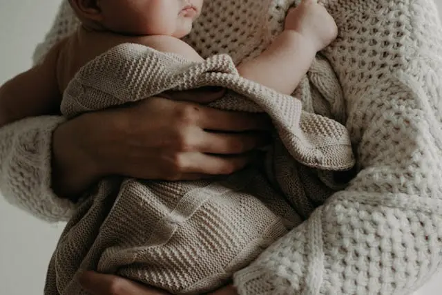 Bébé entouré d'une couverture beige en laine dans les  bras de sa maman avec gilet en laine blanc