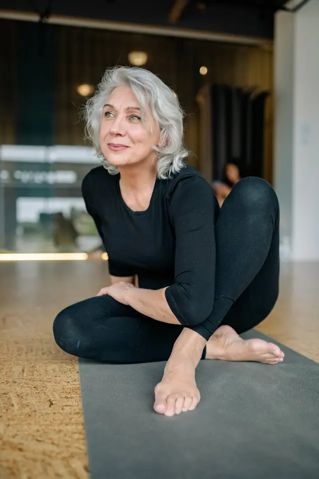 Femme senior cheveux blancs et habits de sport noirs qui sourit  assise les jambes croisées sur un tapis de yoga gris