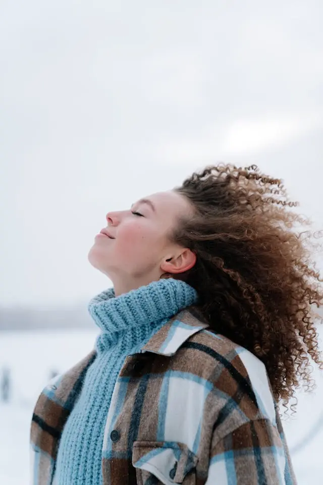 Jeune femme cheveux bouclés avec pull col roulé bleu clair et chemise à carreaux  marron/ blanche et bleu clair qui lève la tête yeux fermés en souriant de profil