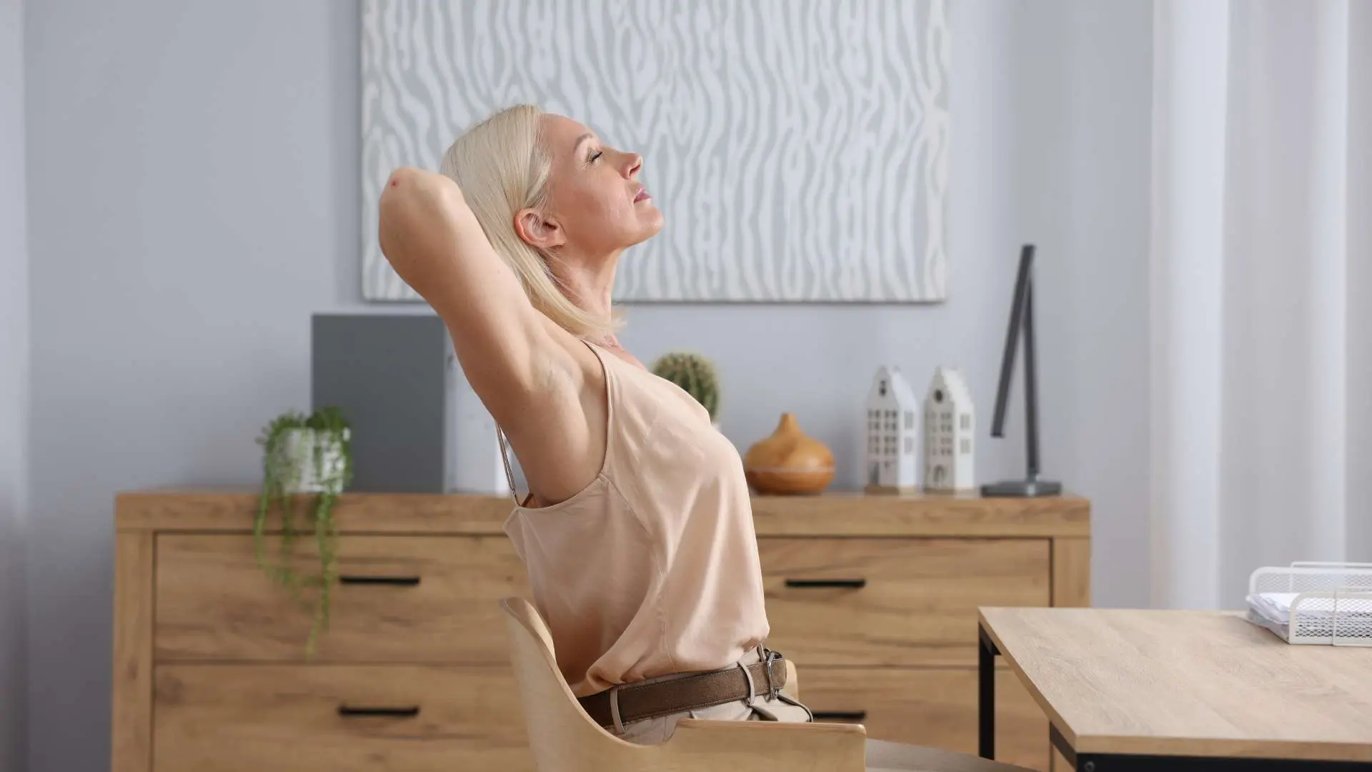Femme de profil assise devant une table qui s'étire  les bras derrière la nuque avec buffet en bois clait et tableau moderne blanc et gris en arrière-plan
