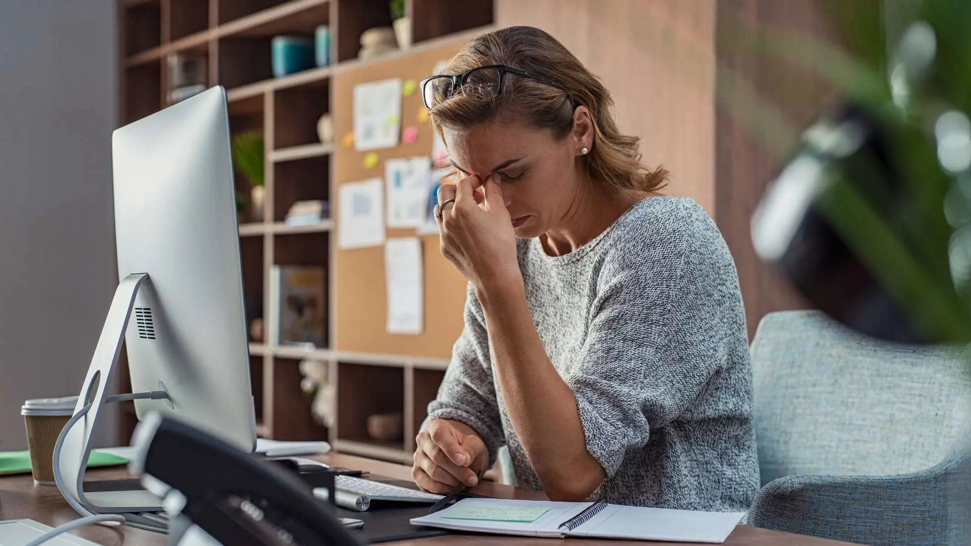 Femme fatiguée assise à son bureau devant ordinateur qui pince le haut de son nez avec sa main gauche la tête baissée