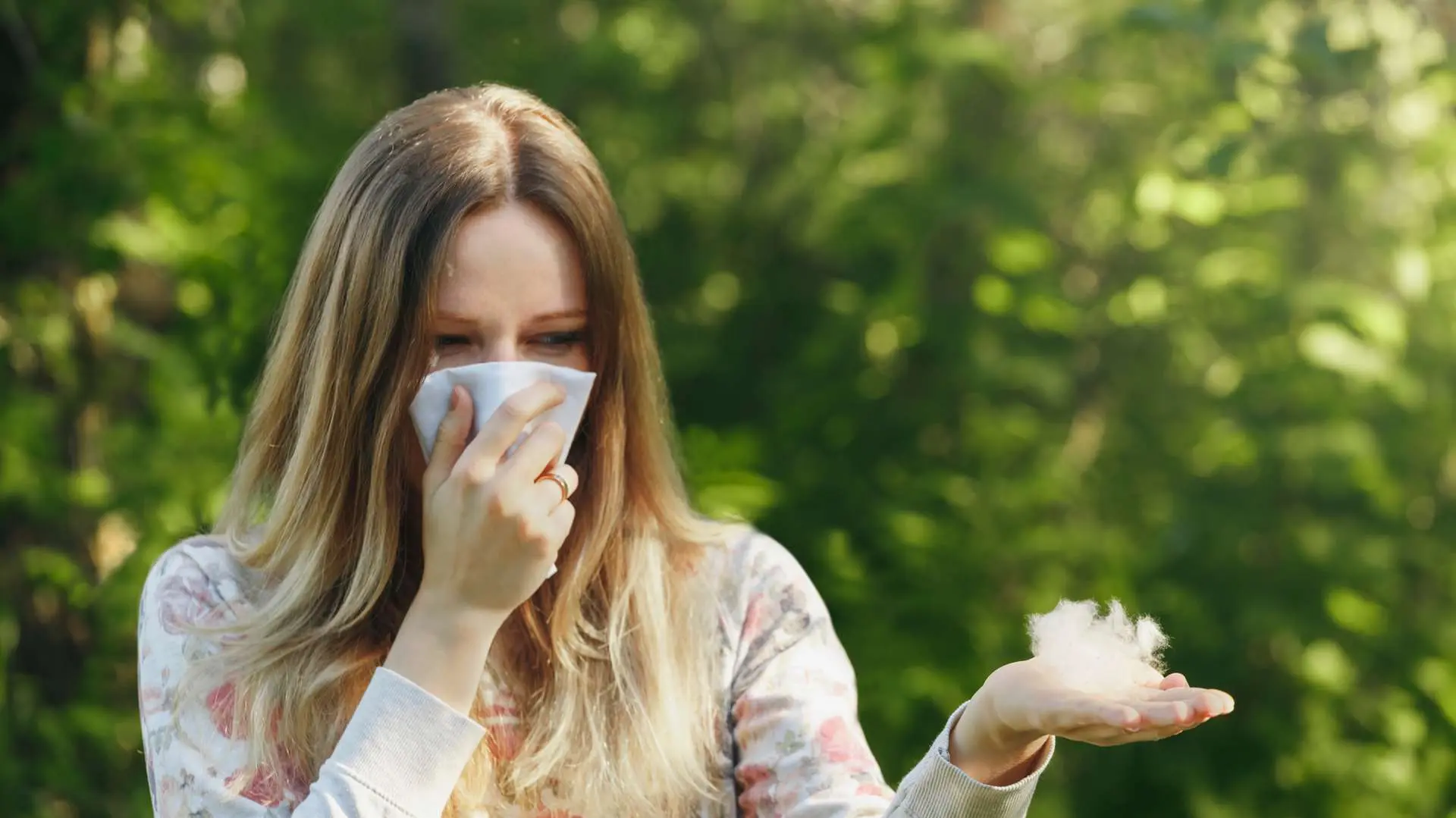 Jeune femme aux cheveux longs avec un mouchoir devant son nez d'une main et l'autre main ouverte avec touffe de fleurs de pissenlit