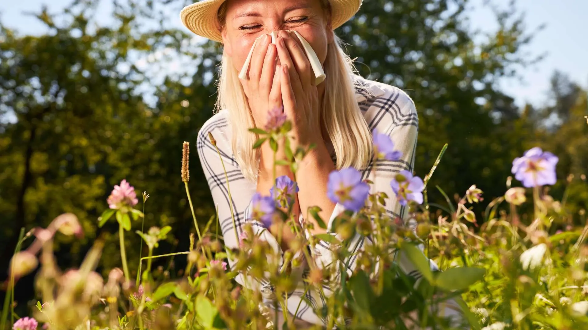 Femme avec chapeau et chemise à carreaux tenant un mouchoir sur son nez devant des fleurs