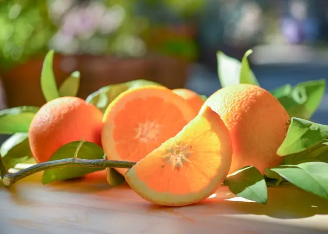 Oranges entières et en quartiers avec feuilles vertes posées sur une table en bois  clair avec rayons de soleil