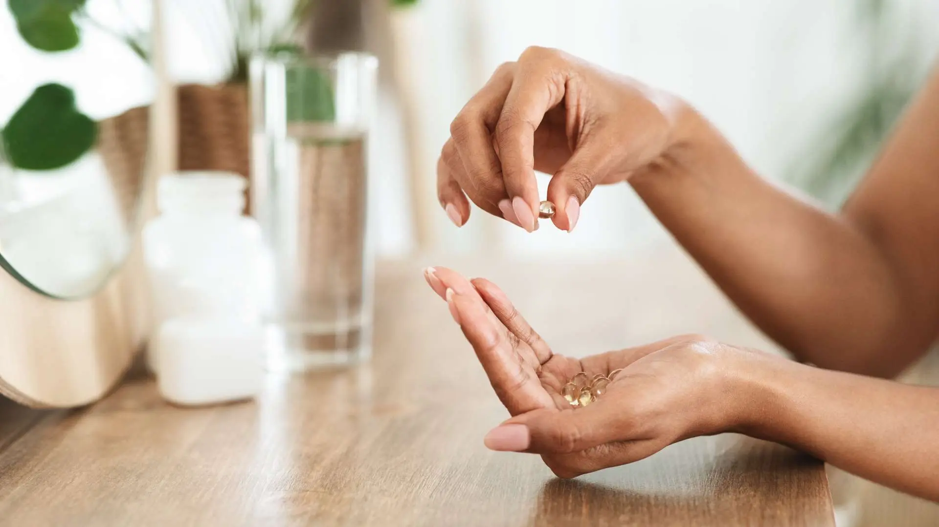 Gros plan main de femme en coup avec compléments alimentaires  et son autre main qui tient un complément alimentaires avec en arrière-plan verre d'eau, flacon blanc et plante posés sur une table en bois clair