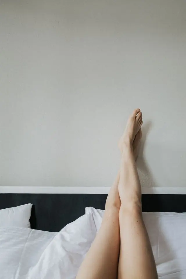 Jambes de femme dans un lit aux draps blancs en l'air contre un mur