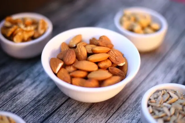 Gros plan ramequin blanc avec amandes dedans entouré de quatre ramequins blancs avec des fruits à coques posés sur une table en bois gris