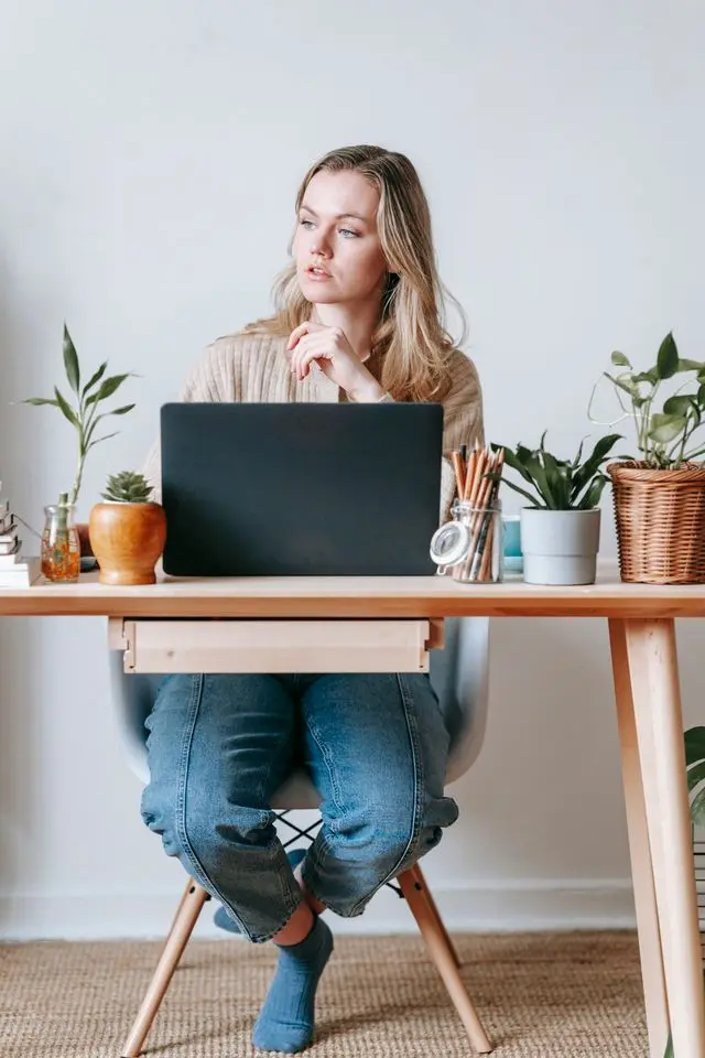 Jeune femme blonde devant ordinateur posé sur bureau en bois avec des plantes qui regarde ay loin pour reposer ses yeux